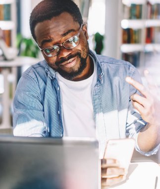 Gözlüklü ve günlük kıyafetleriyle mutlu siyah erkek tasarımcı cep telefonuyla selfie çekiyor ve hafif sanat ofisinde çalışırken zafer jesti yapıyor.
