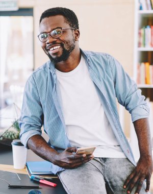 Gözlüklü neşeli Afro-Amerikalı adam rahat bir şekilde dizüstü bilgisayarla masaya oturmuş ve cep telefonuyla sörf yaparken yüzünü çeviriyor.