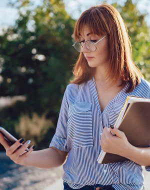 Gözlüklü çekici beyaz kadın sokakta vakit geçirirken cep telefonundan mesaj okudu, düşünceli bayan öğrenci kontrol bildirimi 4G ile bağlantılı 4G kitaplarla şehirde yürüyordu.