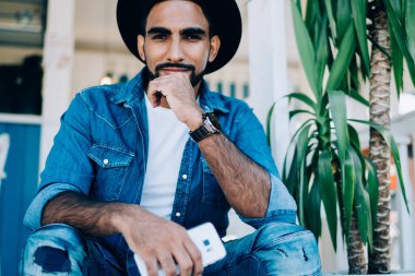 İspanyol asıllı, kendine güveni tam, modaya uygun kot giyen, cep telefonuyla takılan ve kafenin merdivenlerine yaslanmış kameraya bakan bir adamdan.