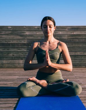 İnce, barışçıl, dişi yogi nilüfer pozisyonunda, elleri bağlı ve gözleri kapalı bir şekilde ahşap kaldırımda oturuyor.