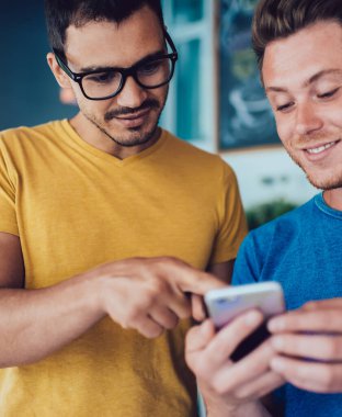 Keyifli genç adam, günlük giysiler içinde, kafeteryadaki akıllı telefon ekranını gösteren gözlüklü yakışıklı arkadaşıyla rahatlıyor.