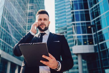 Modern şehir caddesinde güneşli bir günde telefon görüşmesine cevap verirken yüzünü ekşiten ve kameraya bakan sakallı menajerin alçak açısı.