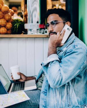 Ciddi etnik erkek, cep telefonuyla bakıcıya sesleniyor ve elinde tek kullanımlık kahveyle caddede alışveriş yaparken internet sorununu çözmeye çalışıyor.