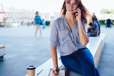 Günlük giysiler ve gözlükler içinde cep telefonu notlarıyla not defteri yazarak sokak kahvesi içerek iş arkadaşıyla proje tartışmakla meşgul.