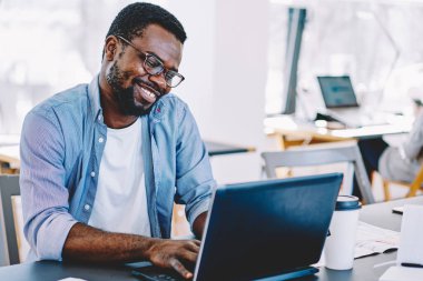 Gözlüklü Afro-Amerikalı adam dizüstü bilgisayarda çalışıyor ve masada büyük pencere ve masaüstlerine karşı otururken mutlu bir şekilde gülümsüyor.