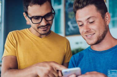 Keyifli genç adam, günlük giysiler içinde, kafeteryadaki akıllı telefon ekranını gösteren gözlüklü yakışıklı arkadaşıyla rahatlıyor.