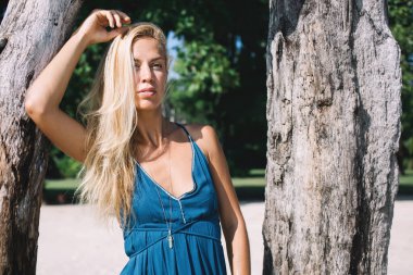 Sarışın, genç, düşünceli, mavi elbiseli bir kadın güneşli bir günde ağaçların arasında dikiliyor ve başka tarafa bakıyor.