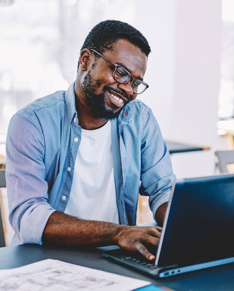 African American man in glasses working at laptop and smiling happily while sitting at table against large windows and desktops