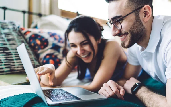 Selective focus side view of smiling couple with trendy haircut having rest on comfy bed while pointing at laptop in modern light bedroom