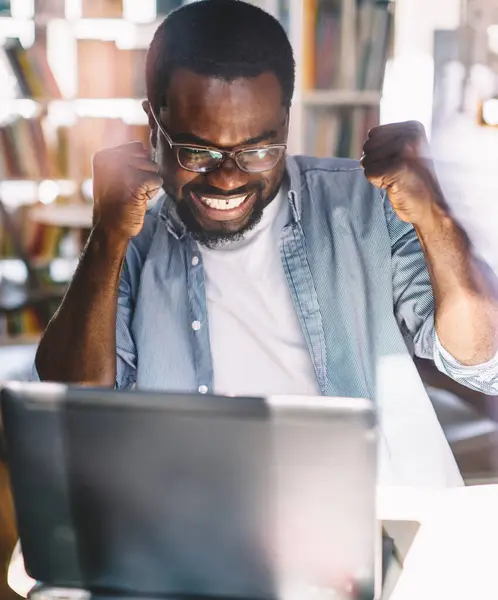 High angle of African American adult overjoyed male in glasses and casual clothes with arms raised celebrating victory while sitting at table and using laptop in light modern office