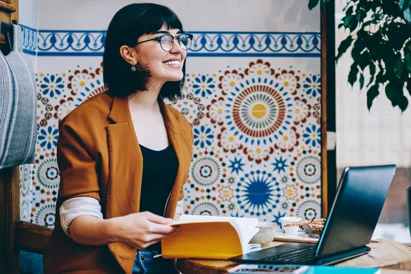 Joyful freelancer in casual wear and glasses using computer working on project reading memo in notebook spending time sitting at table in cafe looking away