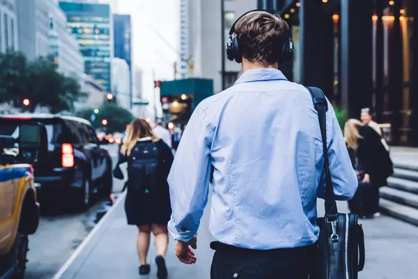Back view of formally dressed intelligent male manager with leather briefcase walking to business meeting in financial district spending way time for listening audio book via electronic headphones