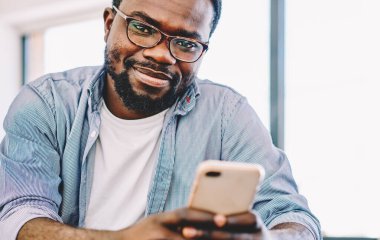 Gözlüklü, gülümseyen sakallı iyimser adam iş arasında kameraya bakarak akıllı telefondan zevkle mesaj atıyor.