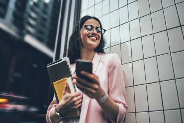 Gözlüklü ve spor ceketli neşeli bir kadının elinde kağıt klasör ve günlükle bina duvarının yanında dururken çok heyecanlı bir şekilde cep telefonu kullanması ve gülümsemesi.