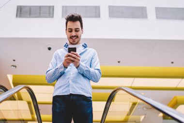 Pozitif neşeli hipster adam yürüyen merdiven üzerinde duran ve merkezinde alışveriş için aşağı gidiyor, mutlu adam boş zamanlarında mobil gadget üzerinde internet tarama cep telefonu yazılımı güncelleme