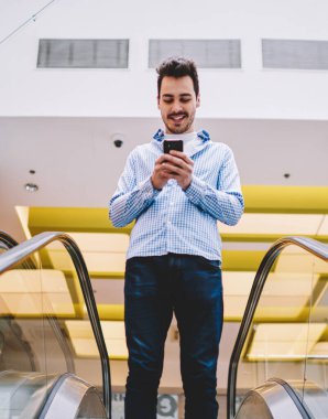 Pozitif neşeli hipster adam yürüyen merdiven üzerinde duran ve merkezinde alışveriş için aşağı gidiyor, mutlu adam boş zamanlarında mobil gadget üzerinde internet tarama cep telefonu yazılımı güncelleme