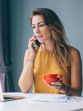 Sarı kolsuz bluzlu özenli genç bir kadın beyaz yuvarlak masada oturuyor elinde laptop kağıdı ve kırmızı bardakla evde akıllı telefondan iletişim kuruyor.