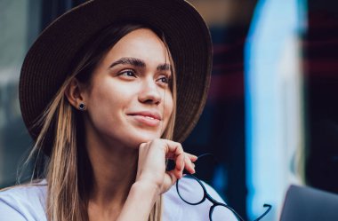 Şapkalı, gözlüklü, kafede otururken tablet kullanan, gülümseyen ve hayal kuran neşeli bir kadın.