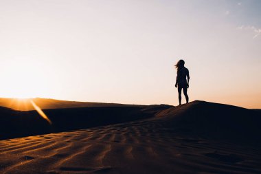 Kadın gezginlerin silueti Fas 'ta tatillerini geçirir ve gün batımında açık gökyüzüne karşı kum tepelerinde yürürler ve ufukta son güneş ışınları belirir