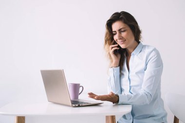 Beyaz tişörtlü yetişkin bir iş kadını cep telefonuyla konuşuyor kahve ve dizüstü bilgisayarla masada oturuyor.