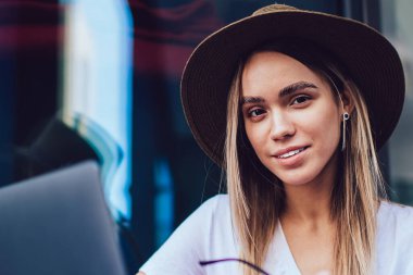 Çekici, kendine güvenen, şapkalı, elinde gözlük olan ve cafe masasında dizüstü bilgisayara bakarken kameraya bakan sarışın kadın.