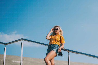 Tatilde, şık sarı saçlı, şık giyimli, güneş gözlüklü, trabzana yaslanmış ve arka planda mavi gökyüzüne bakan bir kadın.