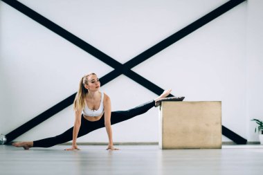 Spor elbiseli, sakin, zayıf, yetişkin bir sporcu gözlerini kaçırıp bacak kaslarını çalıştırırken ön bacak egzersizi yapıyor ahşap kutuya ve yere yaslanıyor ve siyah spor duvar direnç ekipmanlarına yaslanıyor.