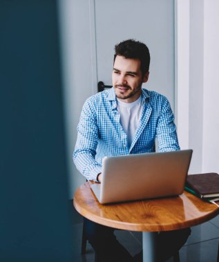 Pozitif düşünceli erkek blogger dizüstü bilgisayar cihazı ile masaüstünde oturan ve bir sonraki yayın için fikir hakkında düşünme, modern netbook e öğrenme mola sırasında uzağa bakarak yetenekli adam düşünme