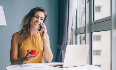 Sarı kolsuz bluzuyla beyaz yuvarlak masada oturan ve evinde akıllı telefondan konuşan kırmızı bardaklı tatmin olmuş genç bir kadın.