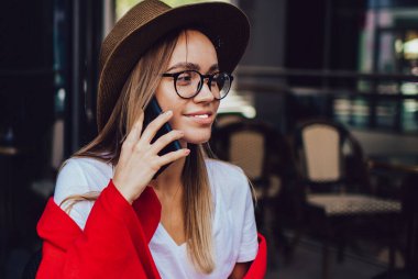 Moda şapkalı ve gözlüklü mutlu bir kadın. Renkli kırmızı battaniyeyle örtülmüş. Akıllı telefonuyla konuşuyor.