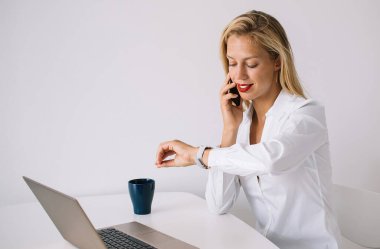 Beyaz masada oturan, bluzlu, gülümseyen genç, sarışın bir kadın. Akıllı telefondan konuşan, dizüstü bilgisayarını kullanan ve saatine bakan.