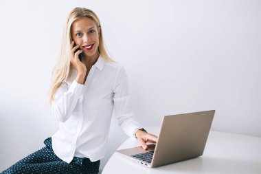 Tatmin olmuş genç sarışın kadın bluzuyla beyaz masada oturuyor akıllı telefonuyla konuşuyor bilgisayarını evde kullanıyor ve kameraya bakıyor.
