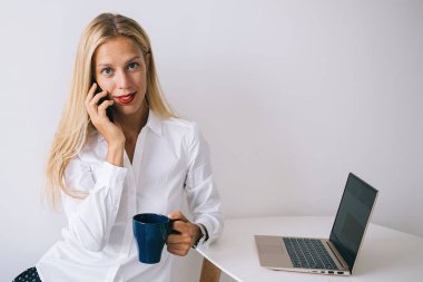Konsantre olmuş mutlu genç sarışın kadın beyaz masaya yaslanmış bluzlu mavi bardakla akıllı telefonla konuşuyor dizüstü bilgisayarını kullanıyor ve beyaz arka plandaki kameraya bakıyor.
