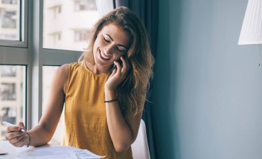 Gözleri kapalı gülümseyen genç bir kadın, günlük giysiler içinde evde akıllı telefondan konuşurken masa başında kağıtlar ve laptopla oturuyor.