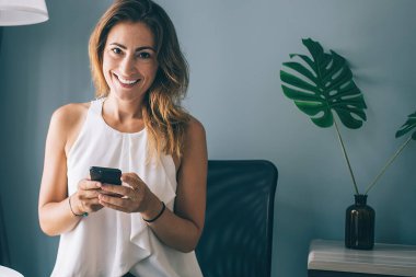 Beyaz kolsuz bluz giyen, akıllı telefondan mesaj atan evde dizüstü bilgisayar kullanan ve kameraya bakan hoş, esmer bir kadın.