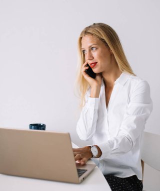 Konsantre olmuş genç sarışın kadın beyaz masada mavi bardaklı bluzuyla oturuyor akıllı telefonuyla konuşuyor evde dizüstü bilgisayarını kullanıyor ve gözünü kaçırıyor.