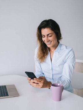 Gömlekli genç esmer, beyaz masada oturuyor, elinde fincan ve laptopla ofisteki akıllı telefondan mesaj atarken gülümsüyor.