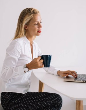 Bardakta içkili, bacak bacak üstüne atmış ve dizüstü bilgisayarda beyaz arka planda yazan konsantre genç bir kadının yan görüntüsü.