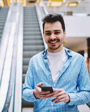 Elinde modern akıllı telefon gadget ile elektrikli merdivenlerde ayakta ve boş zamanlarında iyi hissederken kamera bakarak şık gömlek mutlu gülümseyen hipster adam yarım uzunlukta portre