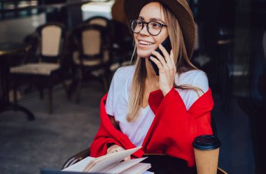 Moda şapkalı, gözlüklü, renkli kırmızı battaniyeli, akıllı telefonlu, dizüstü bilgisayarı ve kahve fincanı olan hoş bir kadın.