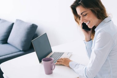 Bardağın ve dizüstü bilgisayarın yanında oturan gülümseyen genç esmerin yan görüntüsü beyaz masada telefonla konuşurken boş bir alan.