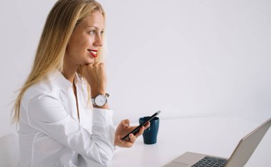 Beyaz masada oturan, mavi bardaklı, beyaz arka planda dizüstü bilgisayar kullanarak akıllı telefondan mesaj atan, konsantre gülümseyen genç sarışın kadının yan görüntüsü.