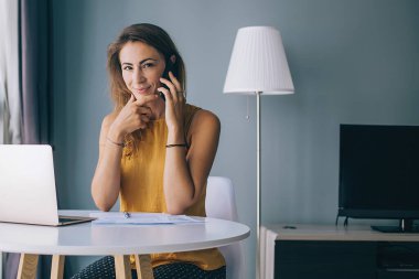 Sarı kolsuz bluzuyla beyaz yuvarlak masada oturan, evinde kameraya bakarak akıllı telefondan iletişim kuran, memnun genç bir kadın.
