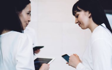 Birbirleriyle konuşurken çeşitli aletler kullanan ve gülümseyen resmi giyinmiş neşeli bayan meslektaşların yan görüntüsü