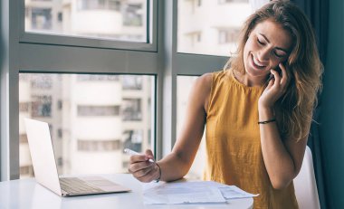 Gözleri kapalı gülümseyen genç bir kadın, günlük giysiler içinde evde akıllı telefondan konuşurken masa başında kağıtlar ve laptopla oturuyor.