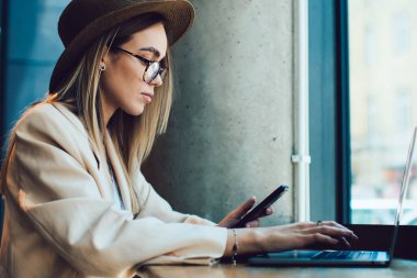 Gözlüklü, şapkalı, elinde telefon olan ve laptopunu modern kafenin penceresinin yanındaki masada oturan zarif yetişkin bayanın yan görüntüsü.