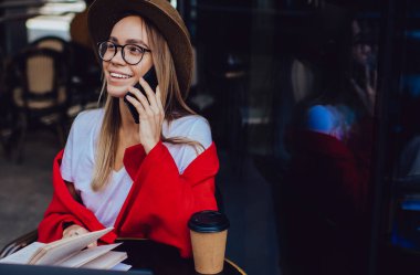 Moda şapkalı, gözlüklü, renkli kırmızı battaniyeli, akıllı telefonlu, dizüstü bilgisayarı ve kahve fincanı olan hoş bir kadın.