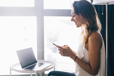 Milenyum kadınlarının gündelik kıyafetlerini giyip evde otururken ve cep telefonuyla mesaj göndermek için interneti kullanırken mutlu kadın blogcu arkadaşlarıyla mobil uygulama üzerinden sohbet ederken yan görünüm.