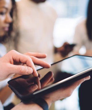 Beyaz tişörtlü bir adamın, iş yerinde çok ırklı insanların yanında dikilirken tablet kullanması.